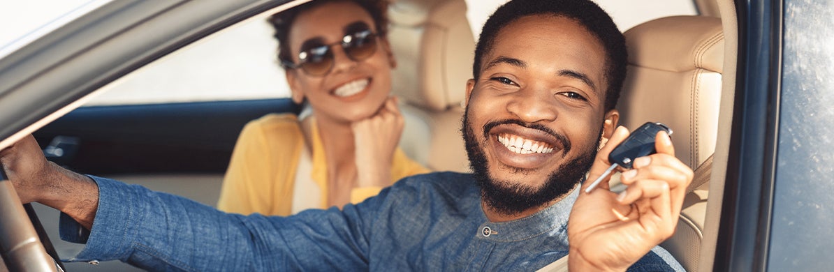 image of a smiling man holding up a car key in the driver's seat with a woman smiling in the passenger's seat in the background
